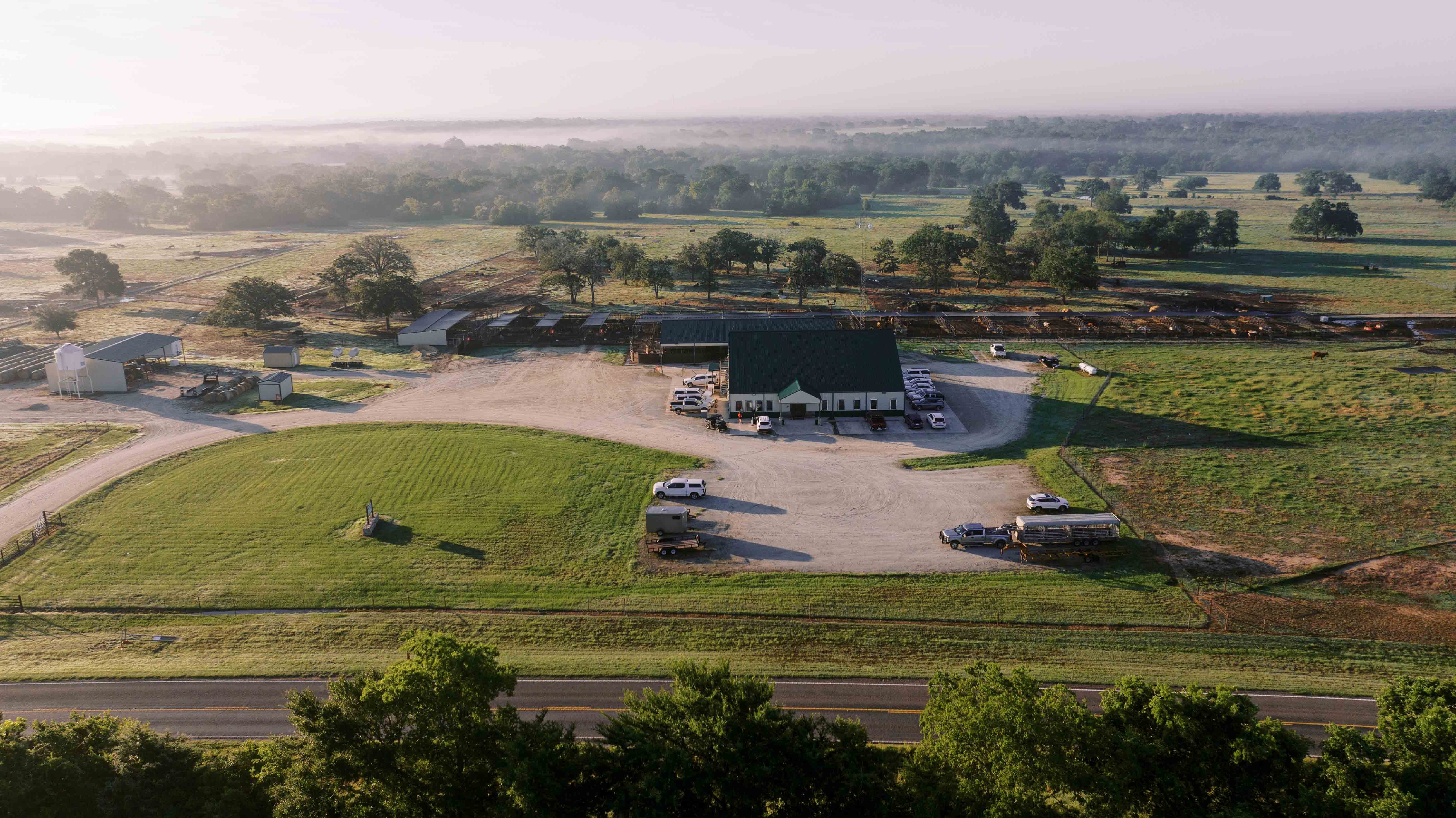 Aerial view of a rural farm with a large building, parked cars, and green fields under a misty morning sky.
