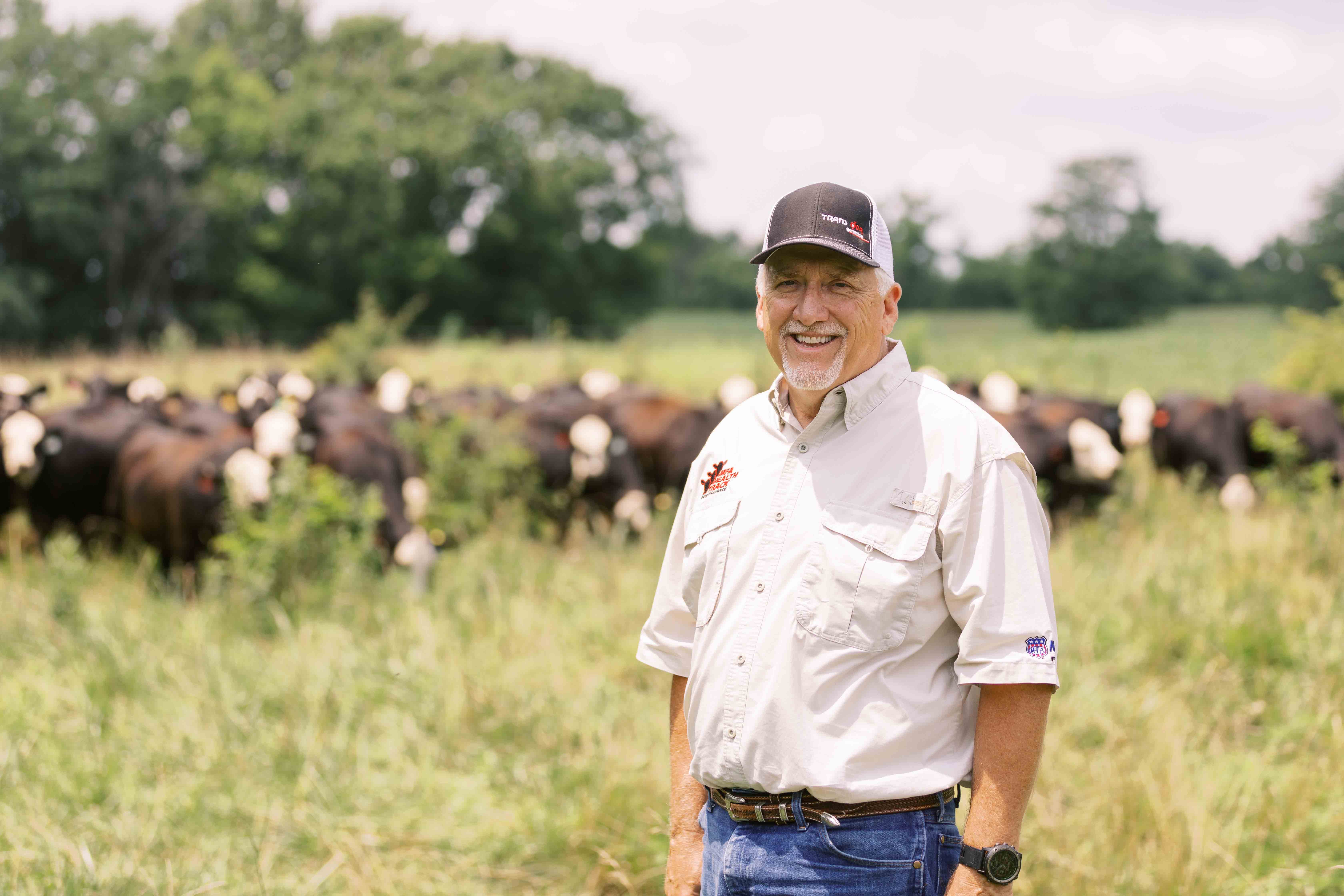 Smiling man in a cap stands in a sunny field with a herd of black and white cows grazing in the blurred background.