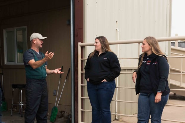 A man in a cap gestures while talking to two women outside a building, with tools leaning against the wall nearby.