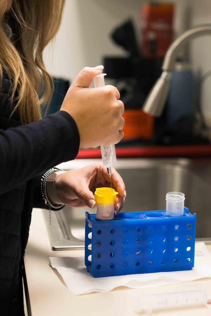 A person uses a syringe to fill a yellow-capped vial in a blue rack, focusing on precise laboratory work at a sink.