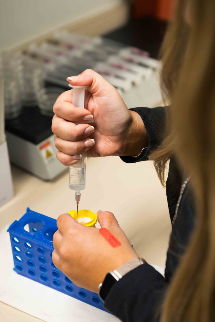 Hands using a syringe to extract liquid from a yellow-capped vial in a lab setting, with a blue rack and blurred background.