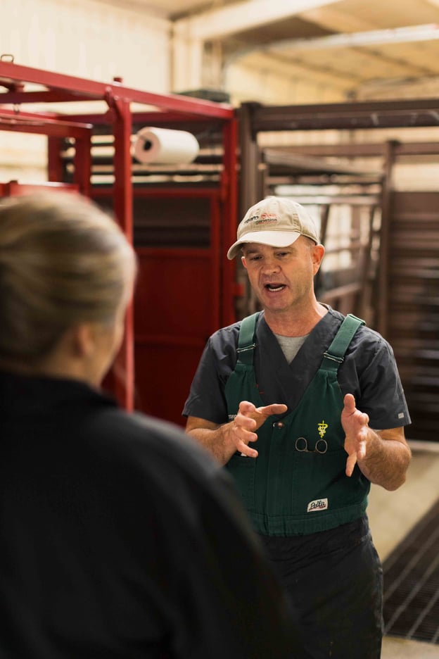 Veterinarian in green overalls explains something to a woman in a barn setting, gesturing with hands, red equipment behind.