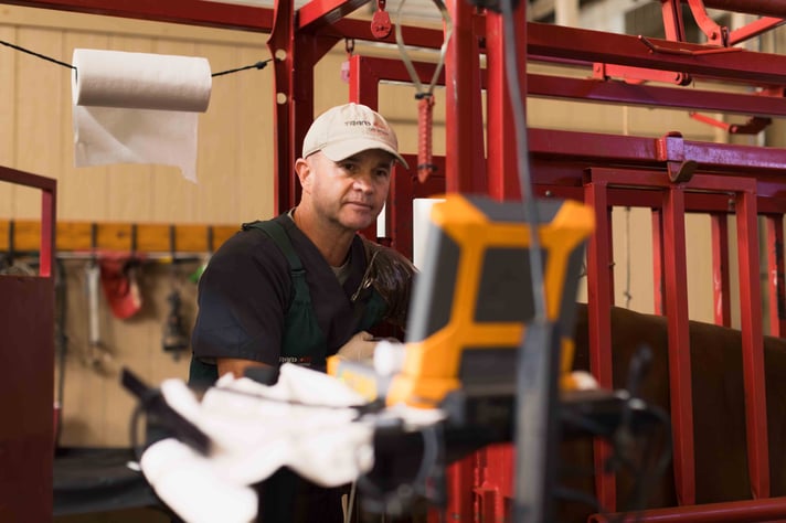 A man in a cap operates equipment in a barn, focused on a screen, with red metal structures and paper roll in the background.