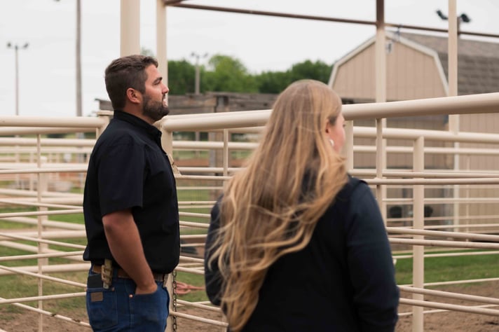 Two people in black shirts stand by a fenced area, observing something in a rural setting with a barn in the background.