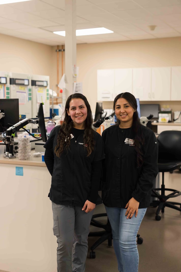 Two genetics laboratory team members standing together in front of lab workstations.